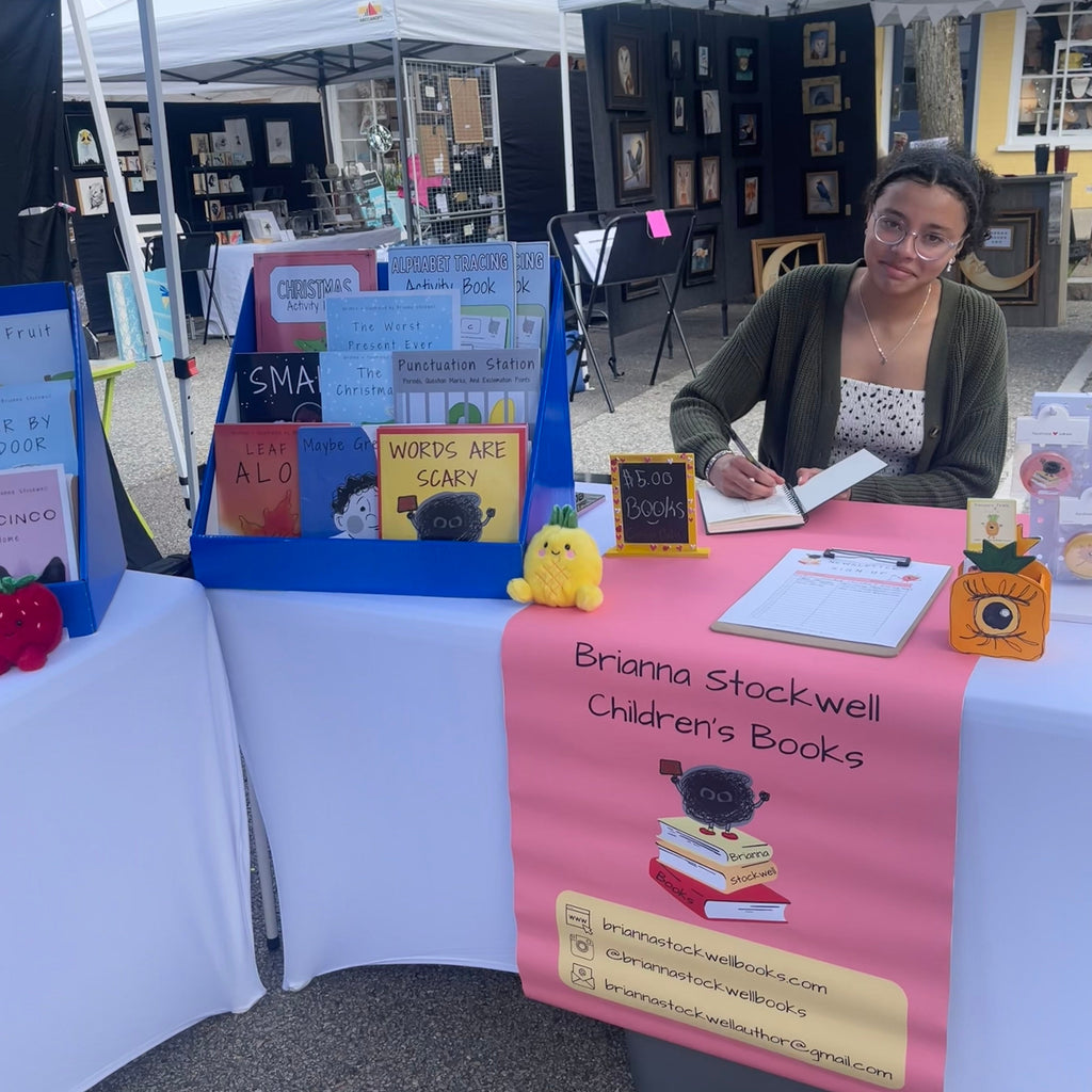 Woman at a book stall with 'Brianna Stockwell Children's Books' sign, surrounded by tents and displays.